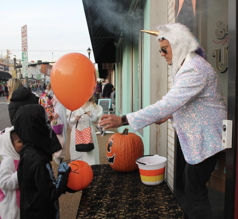 A man dressed up with a unicorn head and a sparkly jacket hands out candy to little kids.