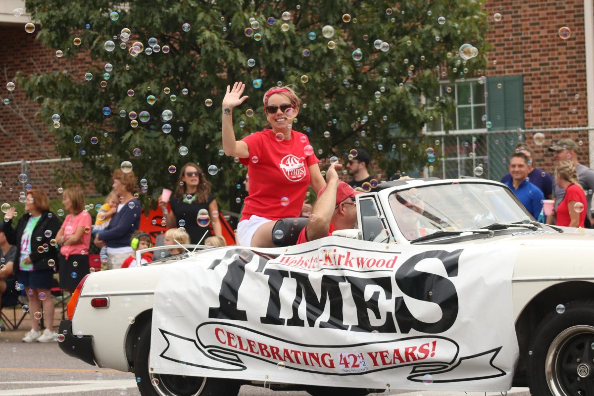 The smiling woman, sitting in the Kirkwood Times newspaper convertible, waves to the people.